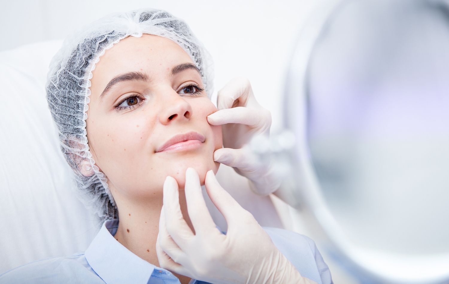 Woman having a cosmetic procedure on her face, being examined by gloved hands in a medical setting.
