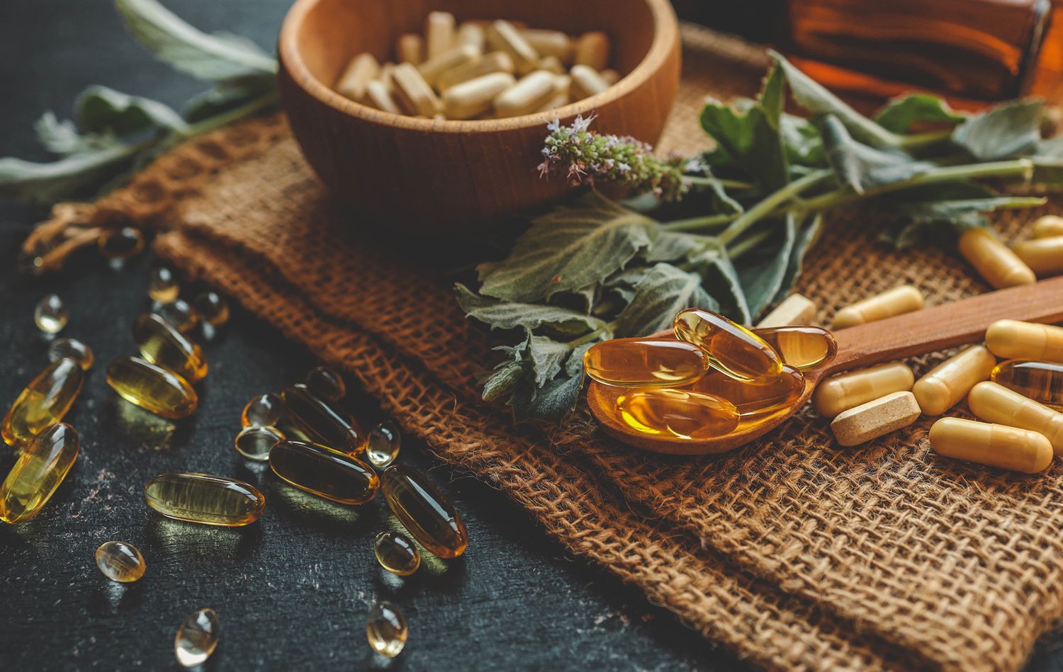 Supplements and herbs on a burlap cloth; includes capsules, a wooden bowl, and a spoon with oil.