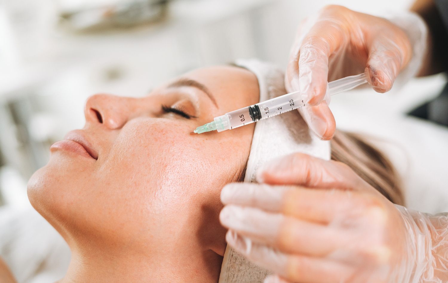 A woman receives a cosmetic injection near her eye, administered by a gloved hand in a medical setting.