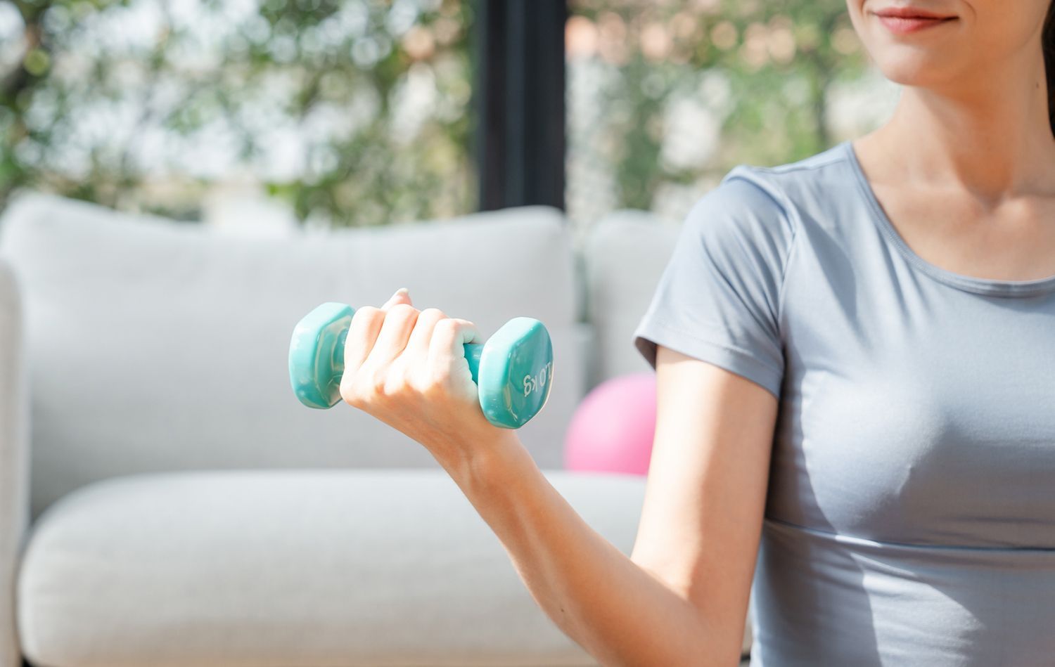 Woman in gray top lifting a teal dumbbell indoors, near a white couch and window.