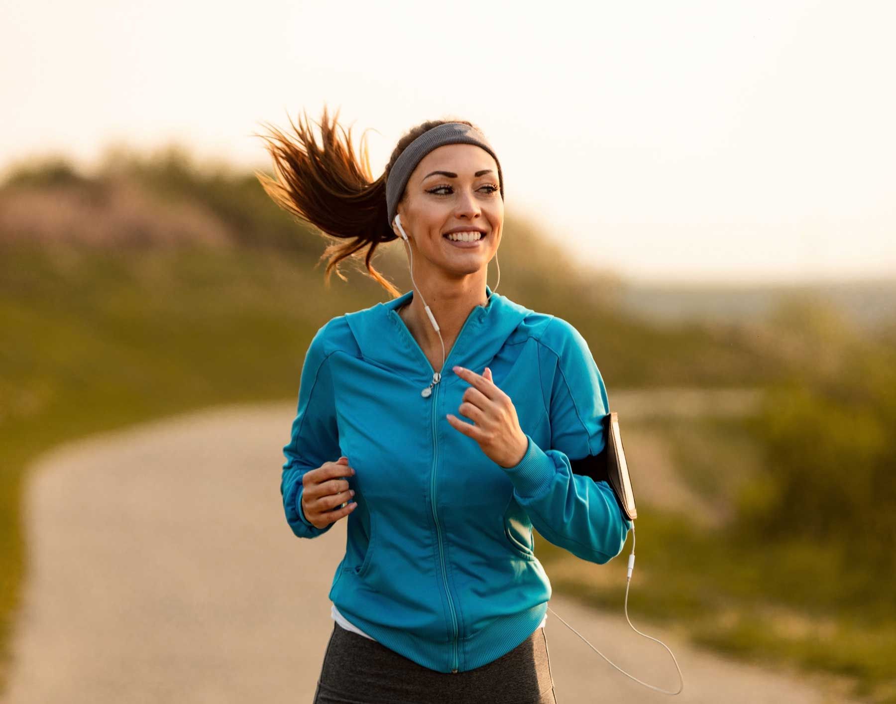Woman running on path, smiling; wearing blue jacket, headband, and armband for phone, with earbuds.