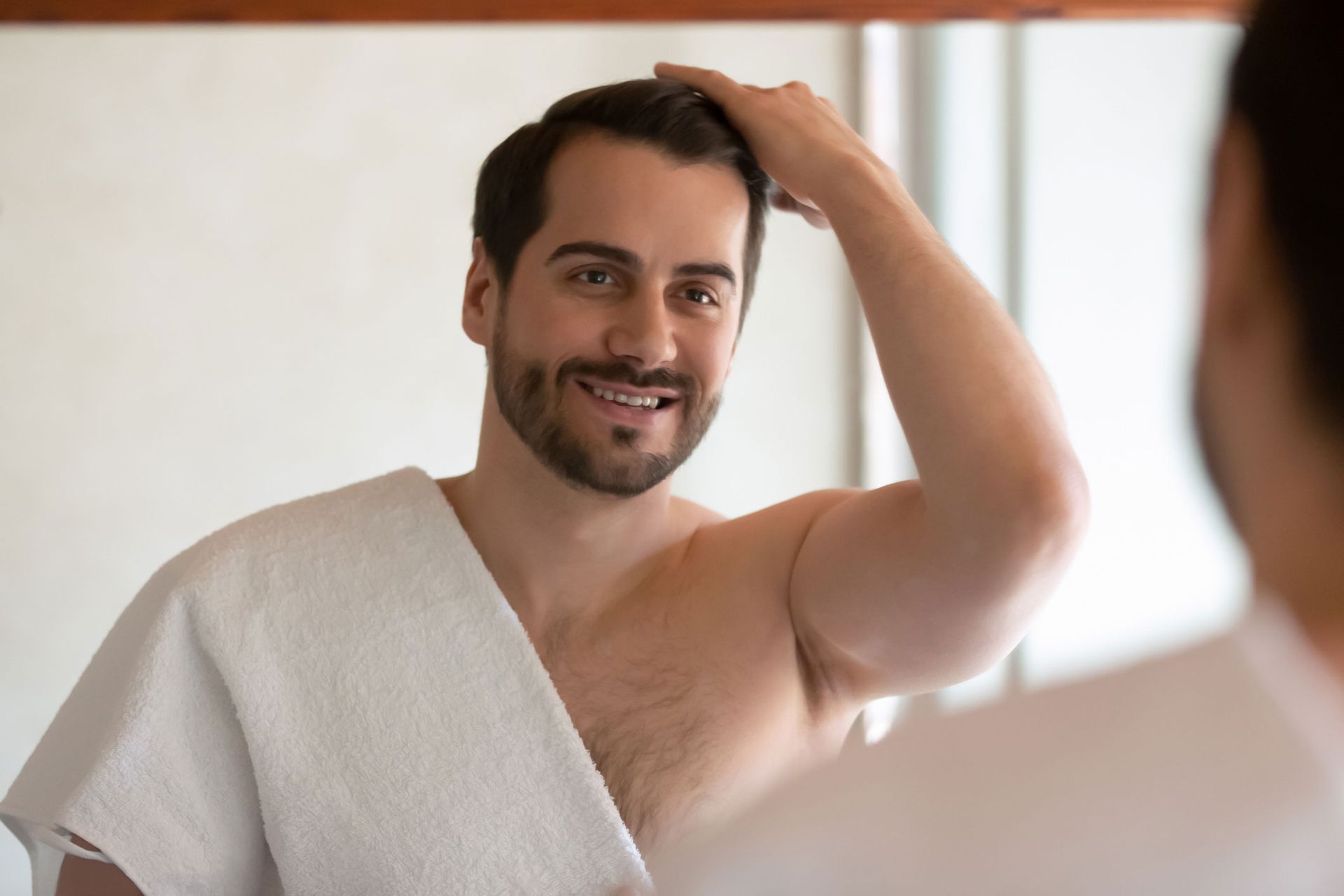 Man in a bathroom, looking in the mirror, smiling, and adjusting his hair with a towel draped over his shoulder.