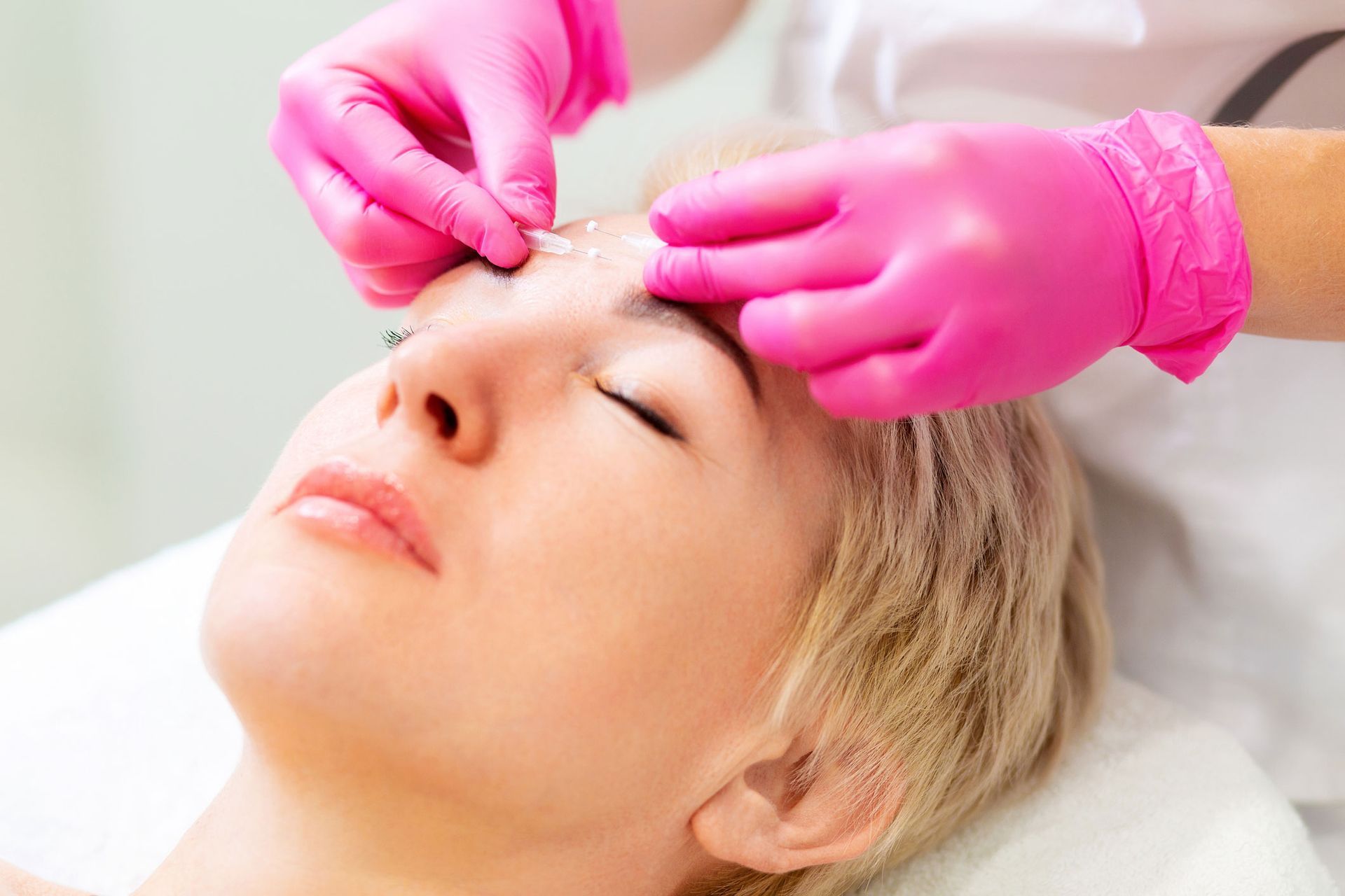Woman receiving facial treatment, pink-gloved hands applying product to her forehead, eyes closed.
