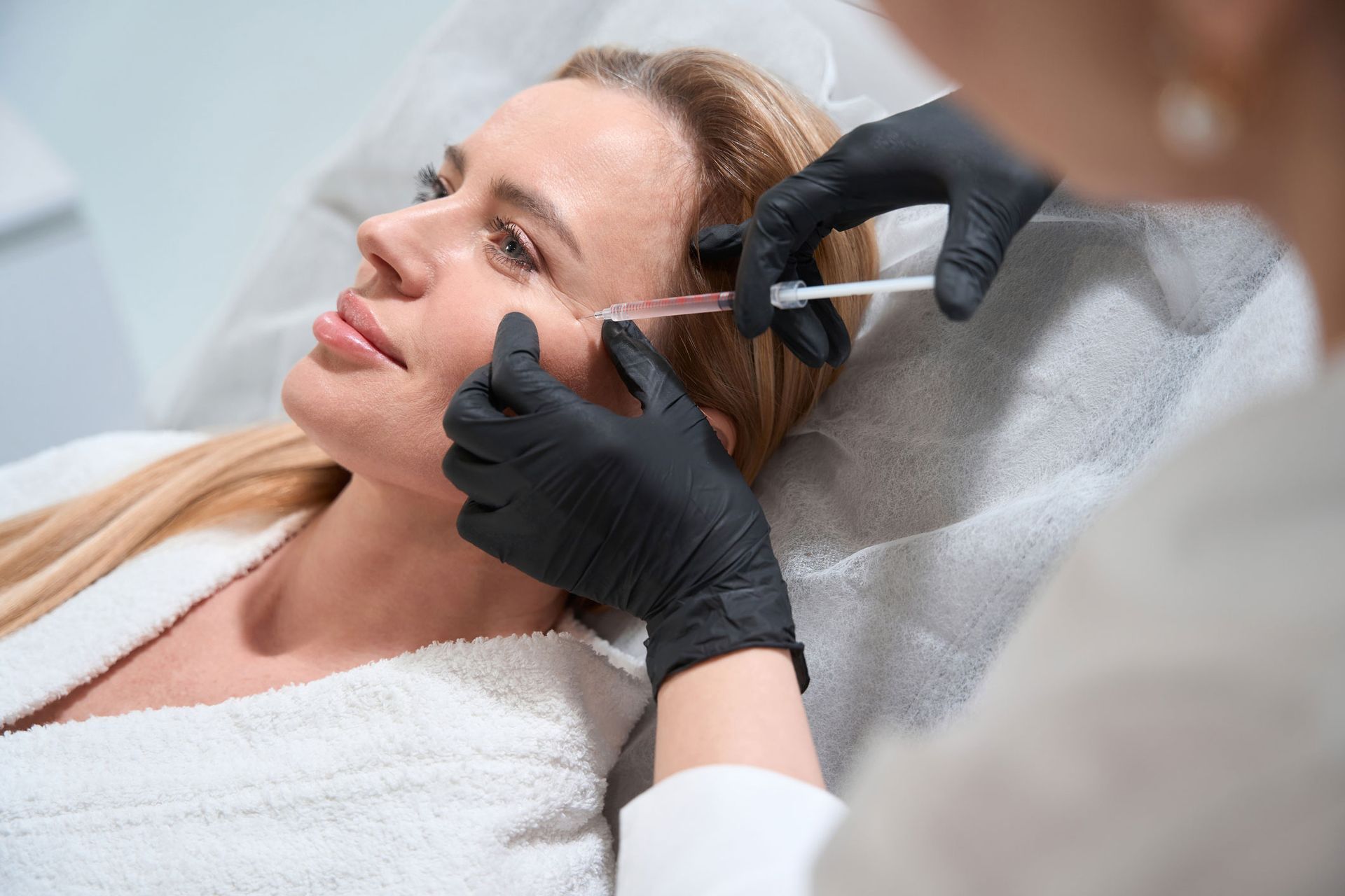 Woman receiving a cosmetic injection near her eye from a person wearing black gloves in a medical setting.