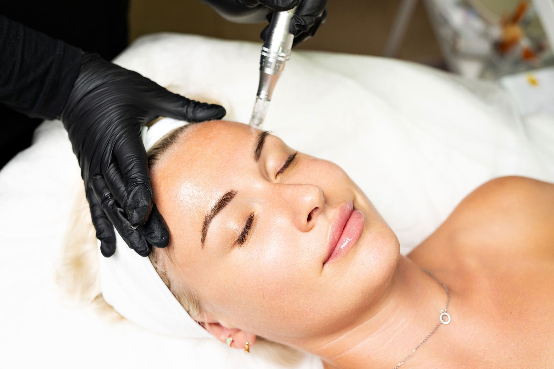 Person receiving a facial treatment; device held near forehead; black gloves; white sheet.