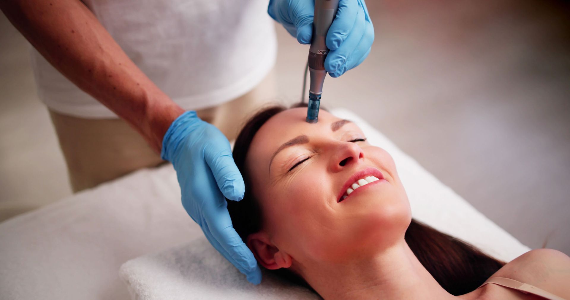 Person receiving facial treatment with tool on forehead. Smiling, light-skinned person in spa setting.